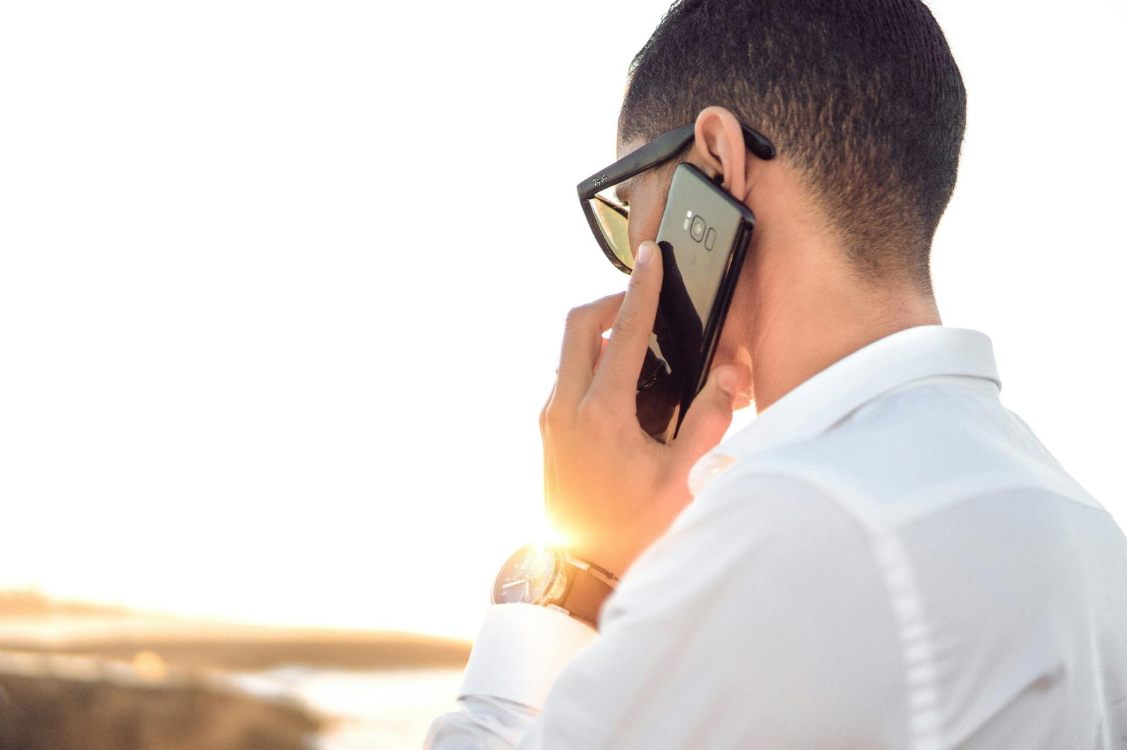 Man outdoors in Morocco talking on smartphone, wearing sunglasses and white shirt, backlit by sunset.